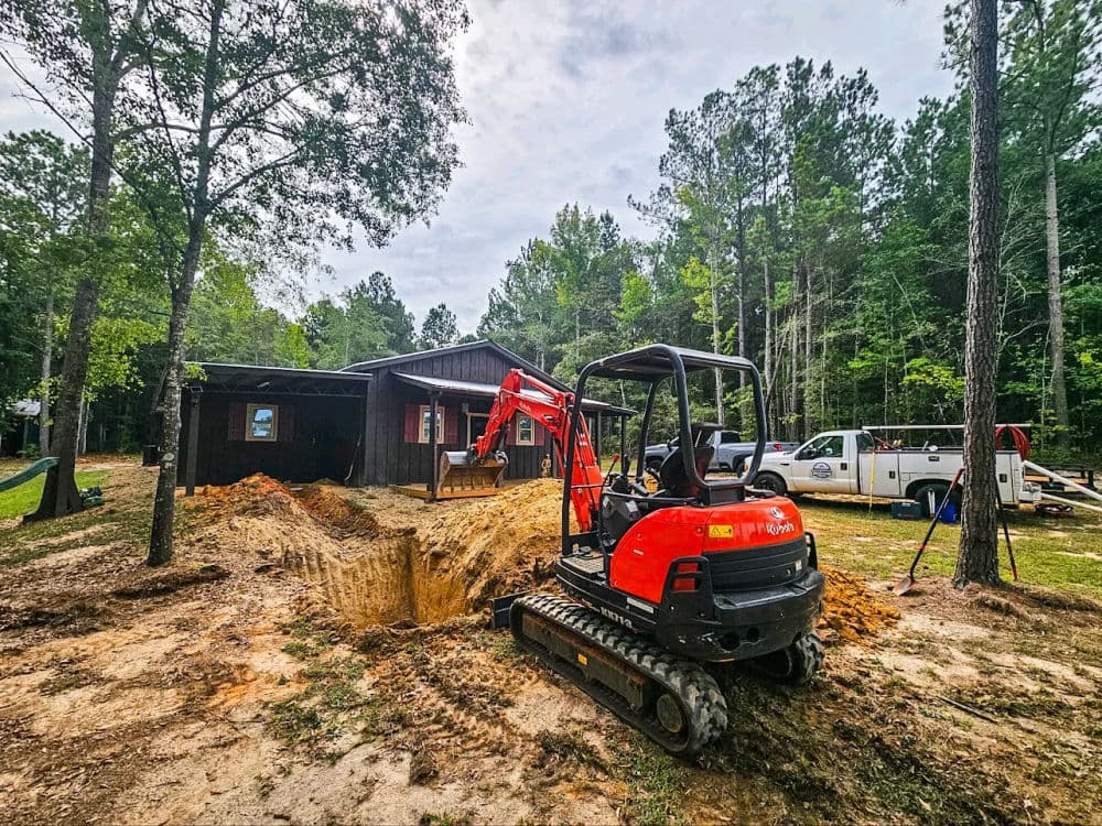 Excavator at residential site preparing for foundation work, surrounded by trees and vehicles.