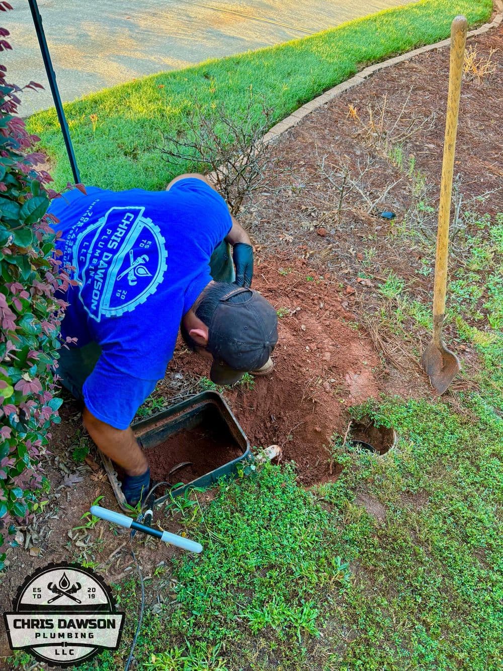 Plumber digging in a garden, repairing a pipe with tools and equipment visible.