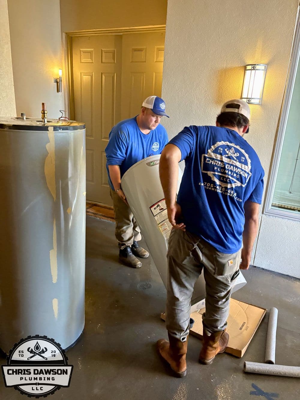 Two plumbers from Chris Dawson Plumbing install a water heater in a residential entryway.