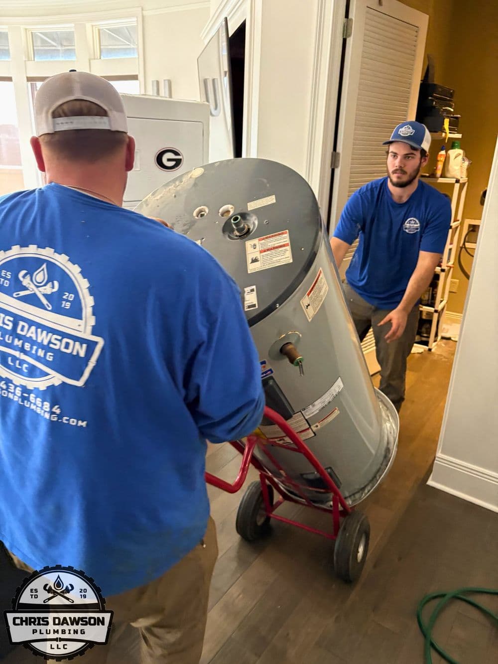 Plumbing professionals moving a water heater with a dolly in a residential setting.