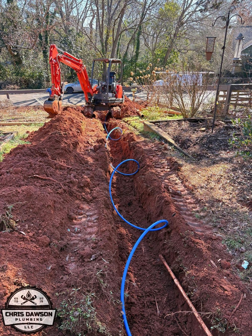 Excavation for plumbing with a backhoe in a residential yard, orange dirt, and blue pipe.