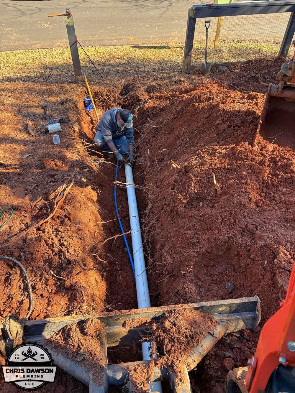 Plumber working in a trench with PVC pipe installation and heavy machinery nearby.