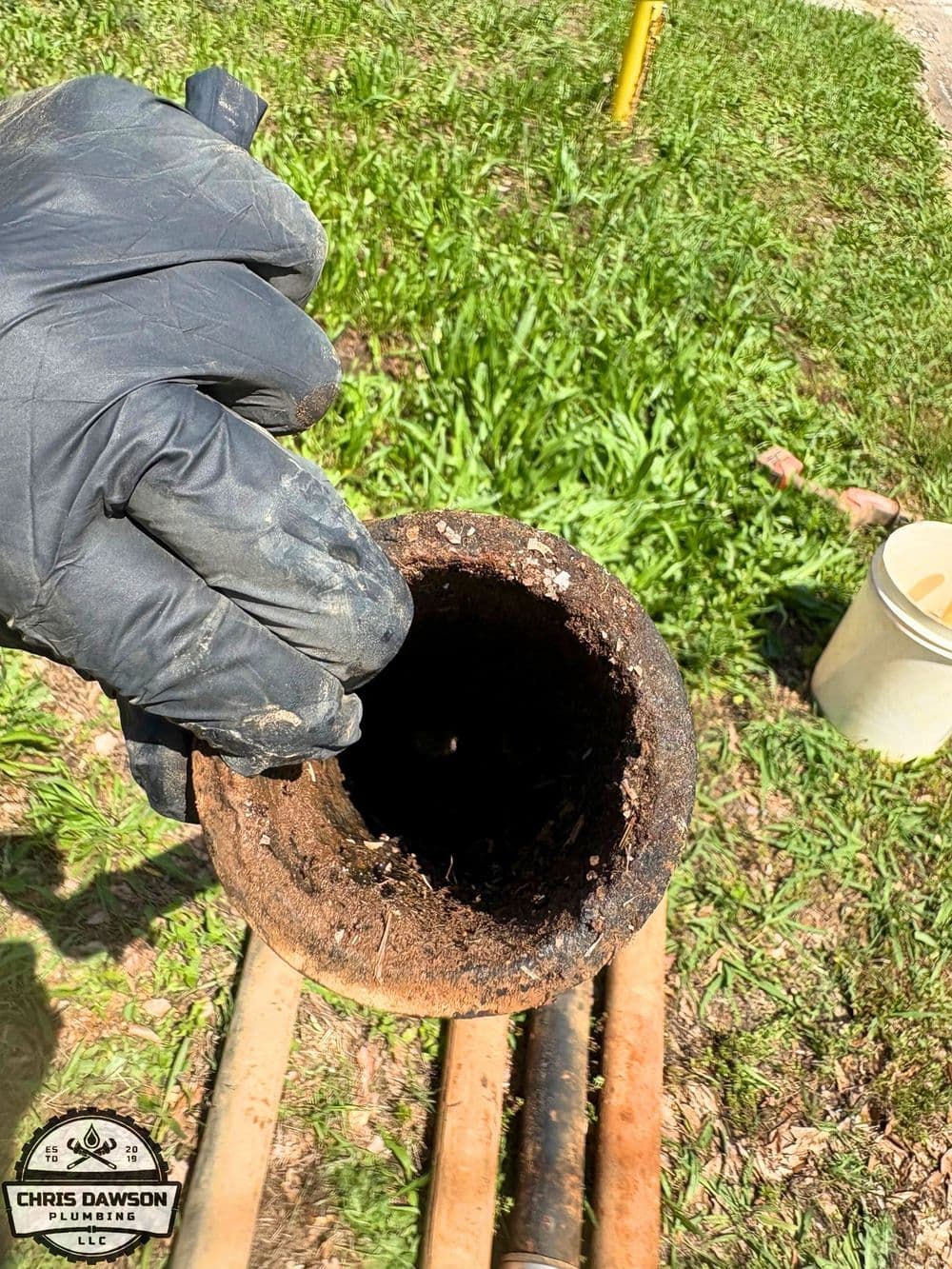 Hand inspecting a rusty pipe showing debris inside, on green grass background.