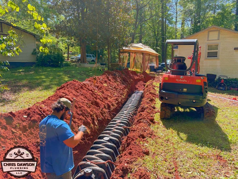 Plumber installing drainage pipe in a trench, with excavator in background and wooded area.
