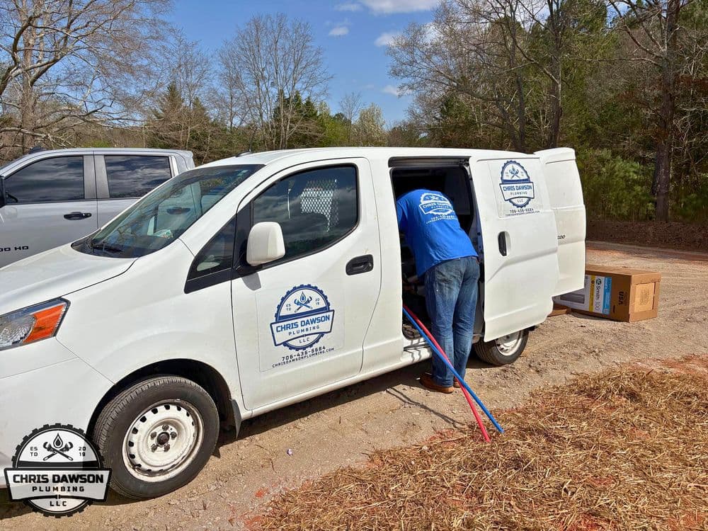 Plumber in blue shirt working from Chris Dawson Plumbing van outdoors in a rural setting.