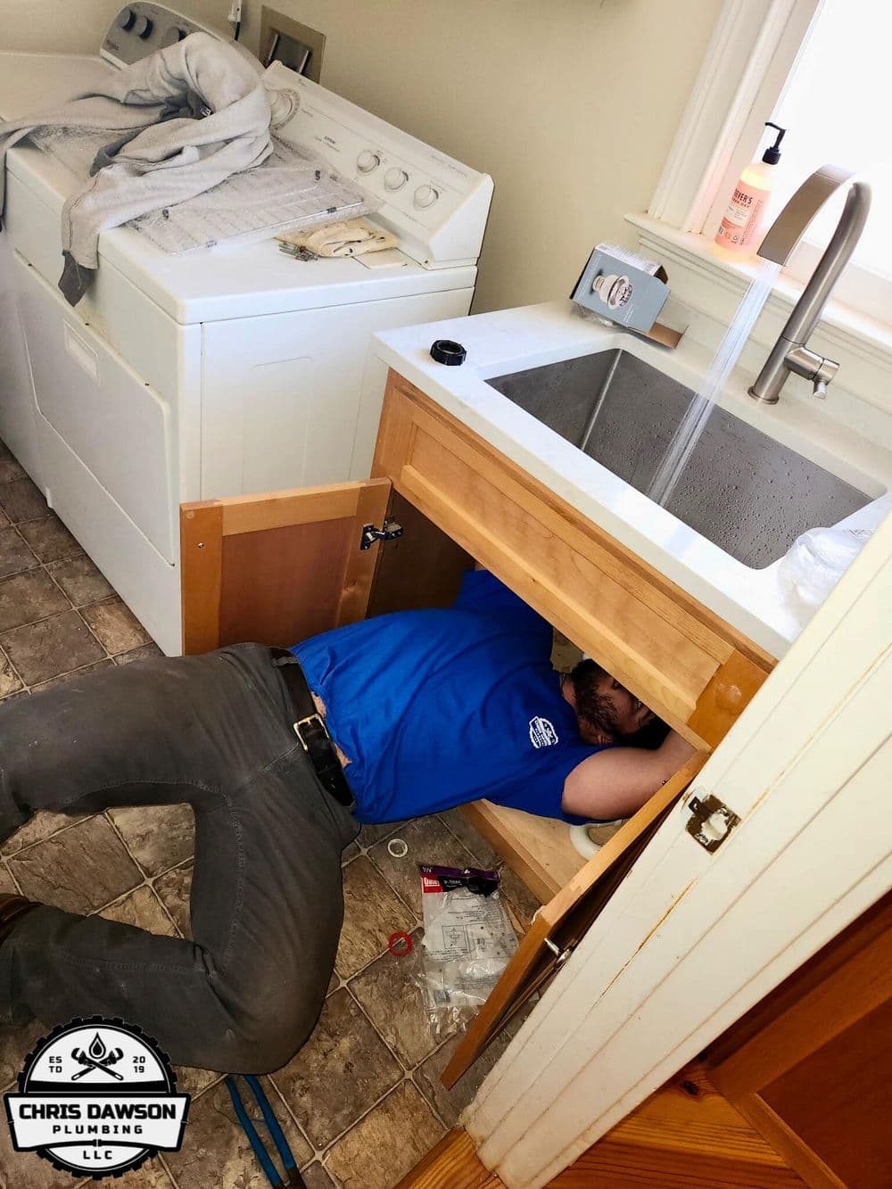 Plumber repairing under-sink plumbing with washing machine in background, Chris Dawson Plumbing logo.