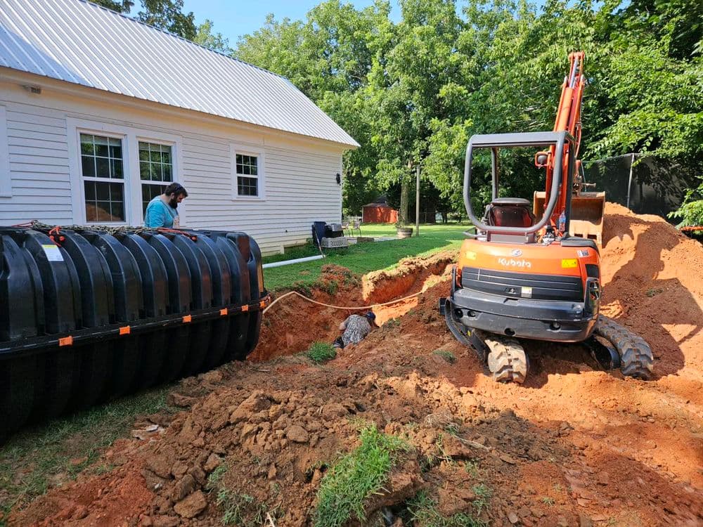 Excavator digging trench for drainage installation beside house with worker present.