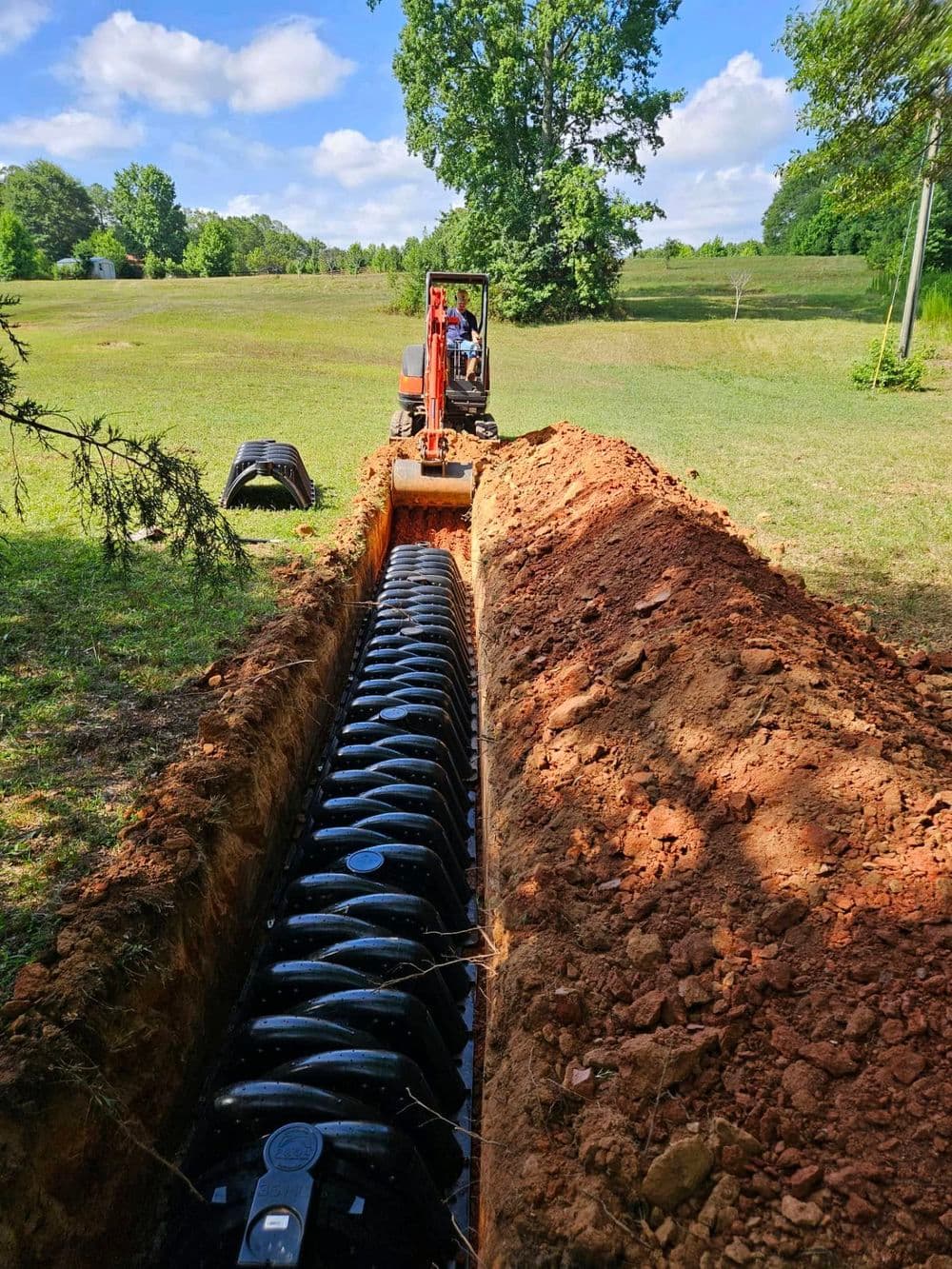 Excavator digging trench for drainage installation in grassy field under blue sky.