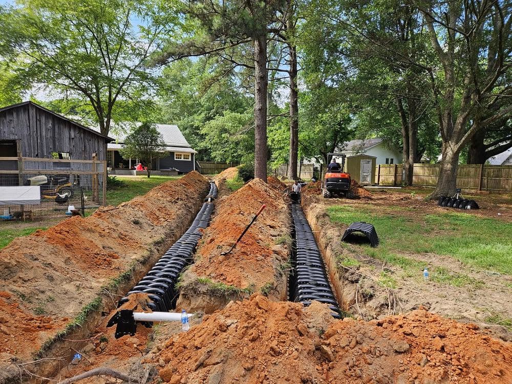 Excavation for drainage system installation in residential yard with machinery and tree cover.